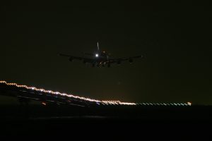Aeronave aproxima-se para pouso em aeroporto durante a noite. É possível notas as luzes do avião e da pista de pouso contra o plano escuro do céu noturno